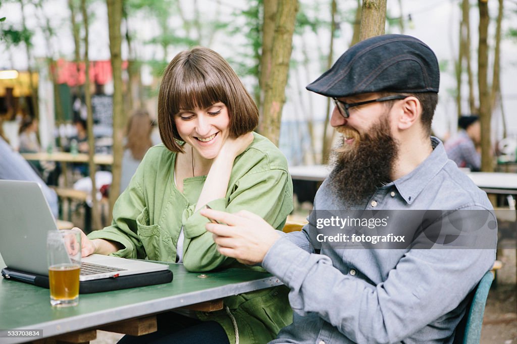 Bearded hipster and girlfriend in beer garden work at laptop