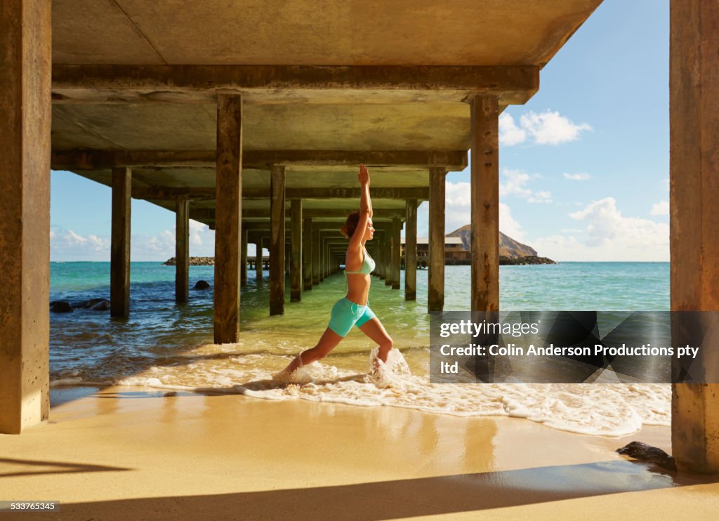 Pacific Islander woman practicing yoga under wooden pier on beach