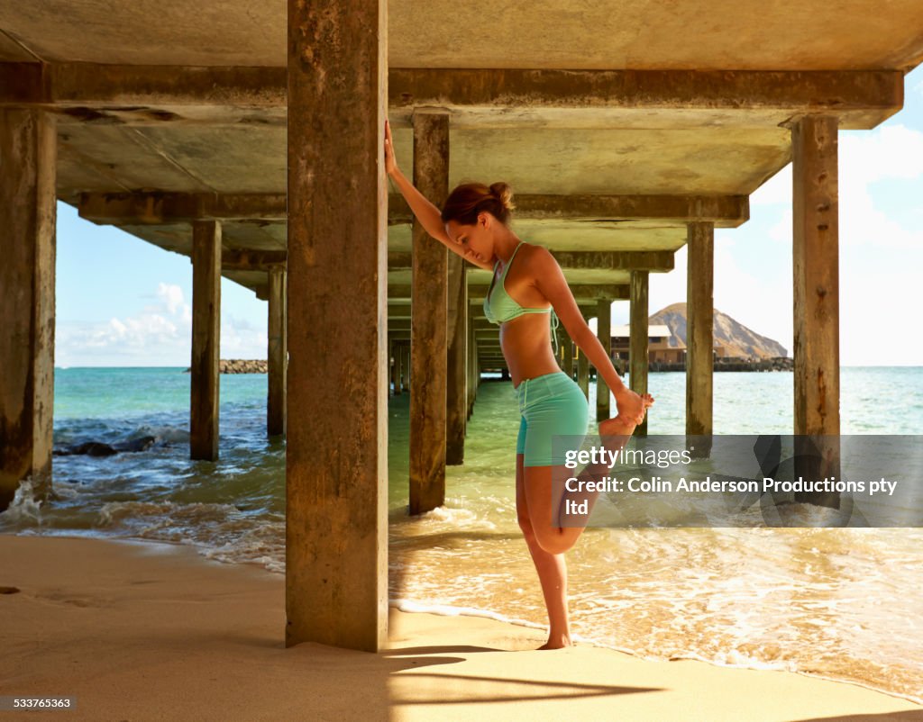 Pacific Islander woman stretching under wooden pier on beach