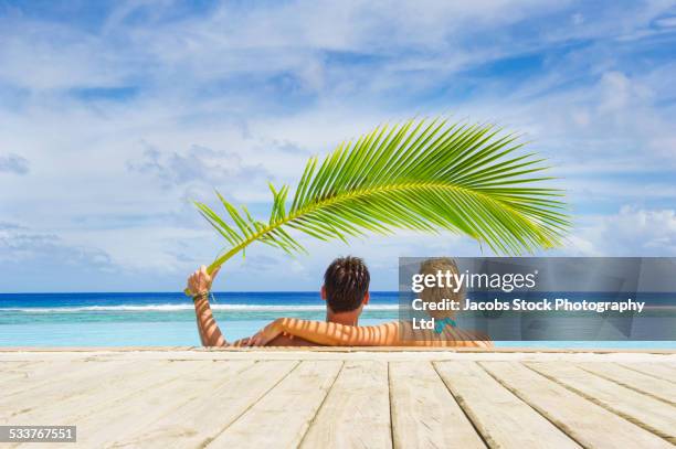 caucasian couple shading themselves under palm frond in swimming pool - palm sunday stockfoto's en -beelden