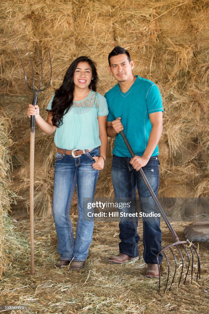 Hispanic couple holding pitchforks near hay in barn