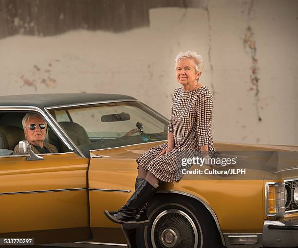 portrait of cool senior woman sitting on bonnet of husbands classic automobile - 69 pose stock-fotos und bilder