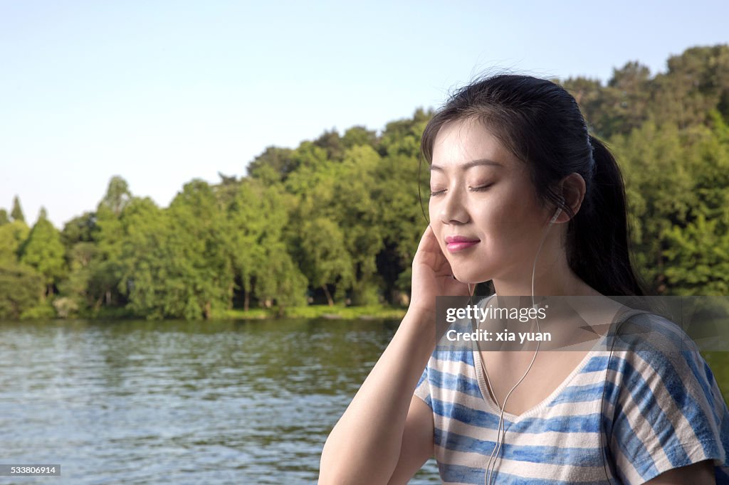 Young asian woman enjoying listening to music on lakeshore,with eyes closed