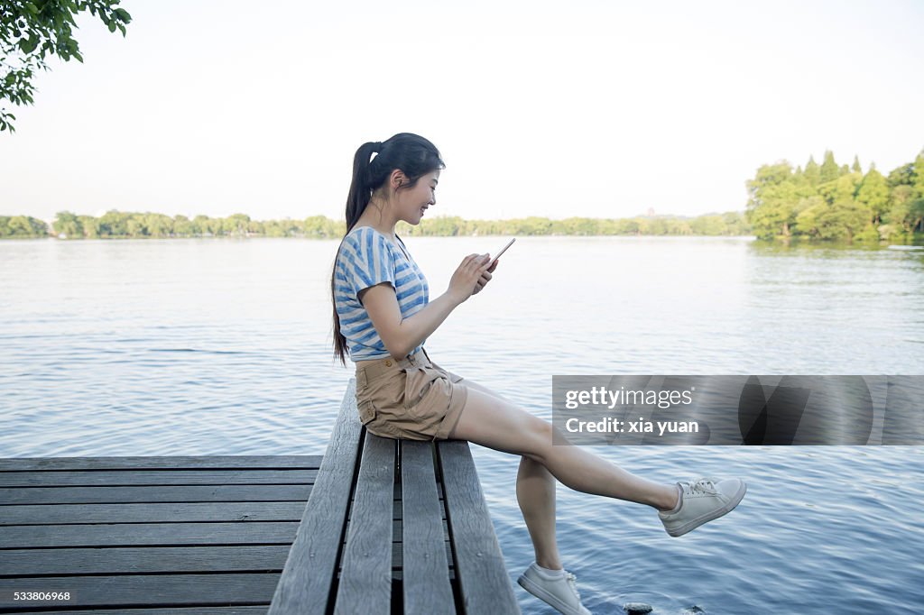 Young asian woman sitting on pier above lake,using smart phone