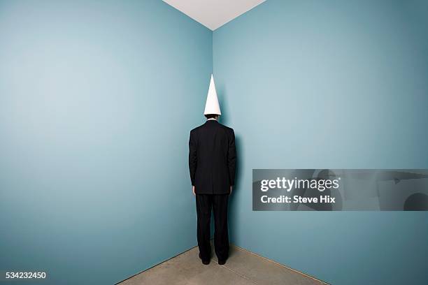 businessman wearing dunce cap standing in corner - dunce cap stock pictures, royalty-free photos & images