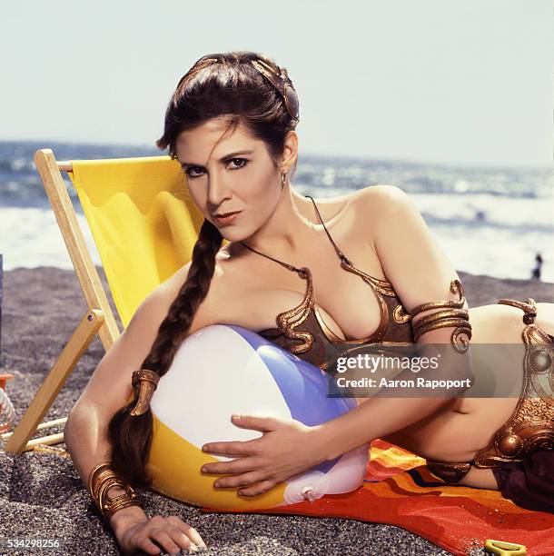 Actress Carrie Fisher as 'Princess Leia' in gold bikini, posing on the beach with an inflatable ball, 1983.