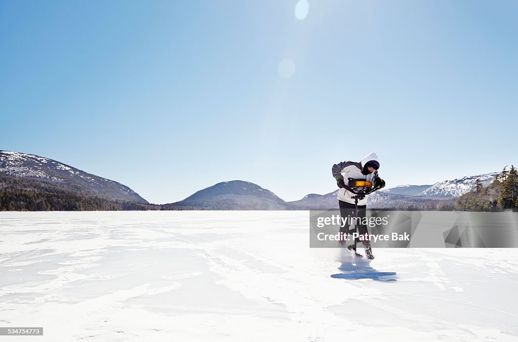 WOMAN ICE FISHING DRILLS ON FROZEN LAKE IN ACADIA