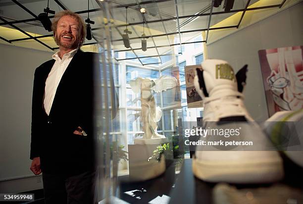 Nike President and CEO Phil Knight standing next to a pair of sneakers on display in a gallery. Knight and Nike helped start a sports business...