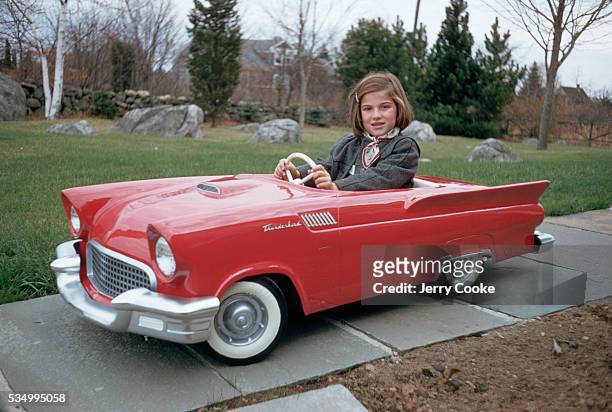 Young girl sits behind the wheel of a child's pedal car version of a Ford Thunderbird.