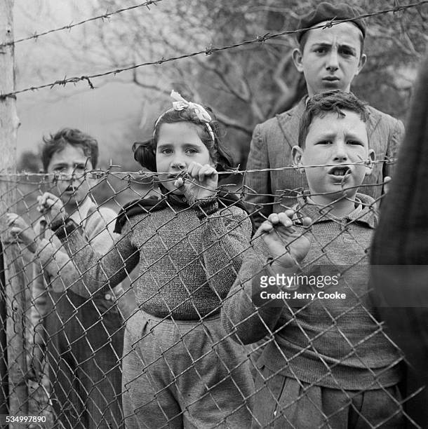 Young refugee from eastern Europe stand behind a chain link fence at a displaced persons camp.