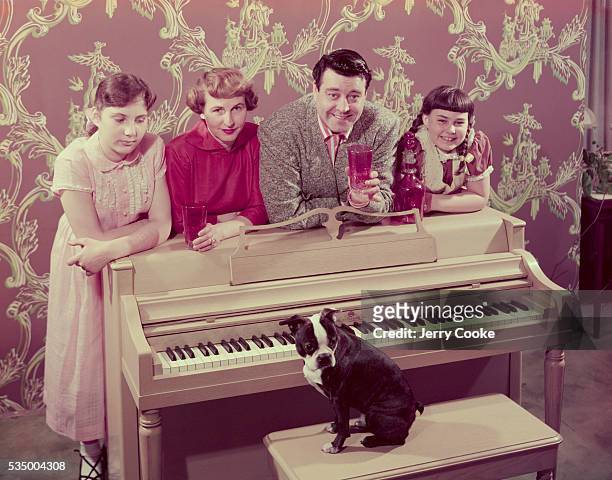 Comedian Jackie Gleason poses at the piano with his wife Genevieve, daughters Linda and Geraldine, and their Boston terrier.