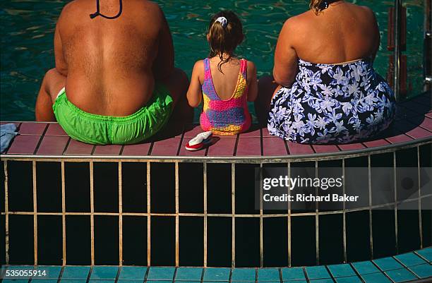 Two parents and their daughter sit with their backs to the viewer on the edge of a pool on the open deck of the Fun Ship Ecstasy belonging to...