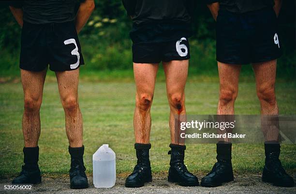 Three soldier recruits wearing shorts and black army boots, one with blood trickling down from the knees to the shins, stand at ease, lined up for...