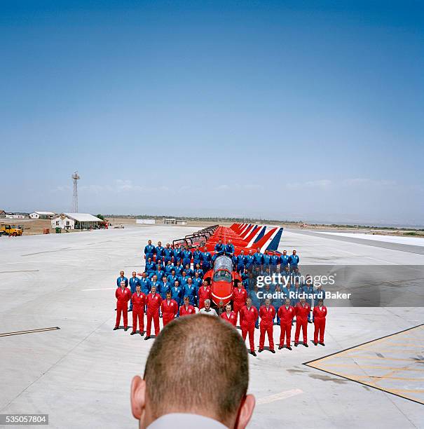 In the mid-day heat, all members of the elite 'Red Arrows', Britain's prestigious Royal Air Force aerobatic team, stand at ease and we see the back...