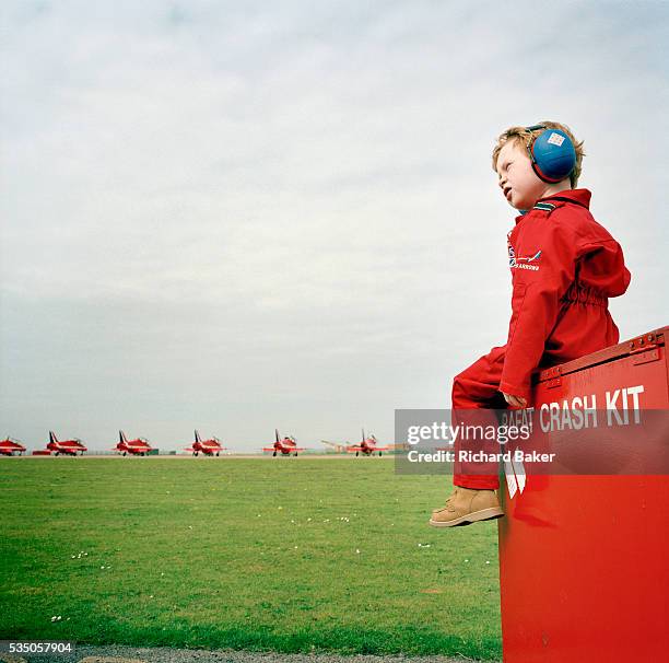 Young boy wearing ear-defenders and dressed in a child's miniature red flying suit is perched on a metal crate of flight spares belonging to the...