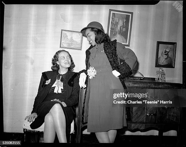 June Eckstine wearing coat trimmed in Persian lamb standing next to seated woman, in interior with buffet and pictures on wall in background,...