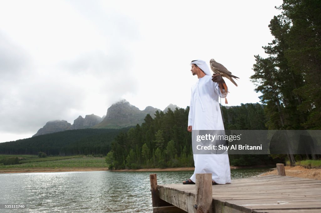 Arab man standing on jetty by lake holding eagle on hand.