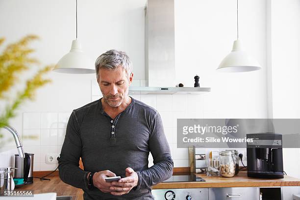 portrait of man standing in kitchen and using smartphone - solo un uomo maturo foto e immagini stock