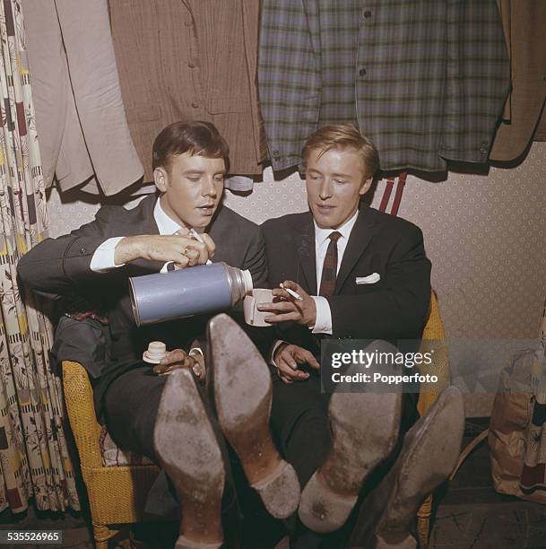 English singers, Mike Sarne and Marty Wilde share a cigarette and hot drink from a thermos flask backstage in their dressing room before a concert...