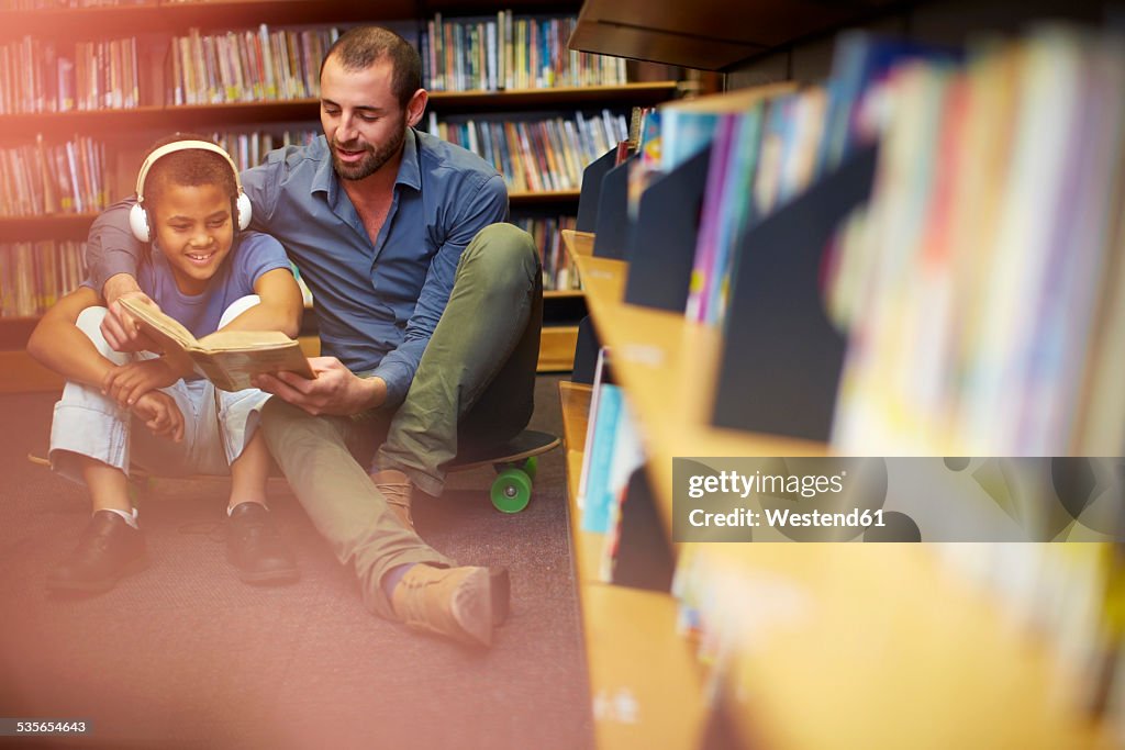 Man reading book to boy with headphones in library