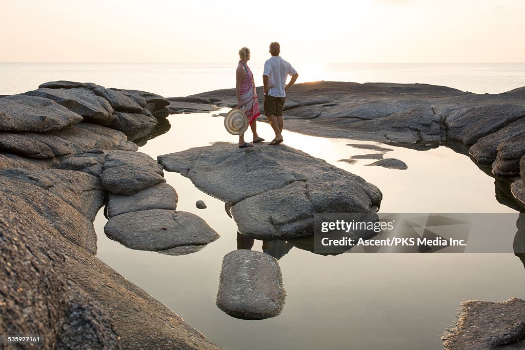 Couple stand on rock island, look out to sea, sun
