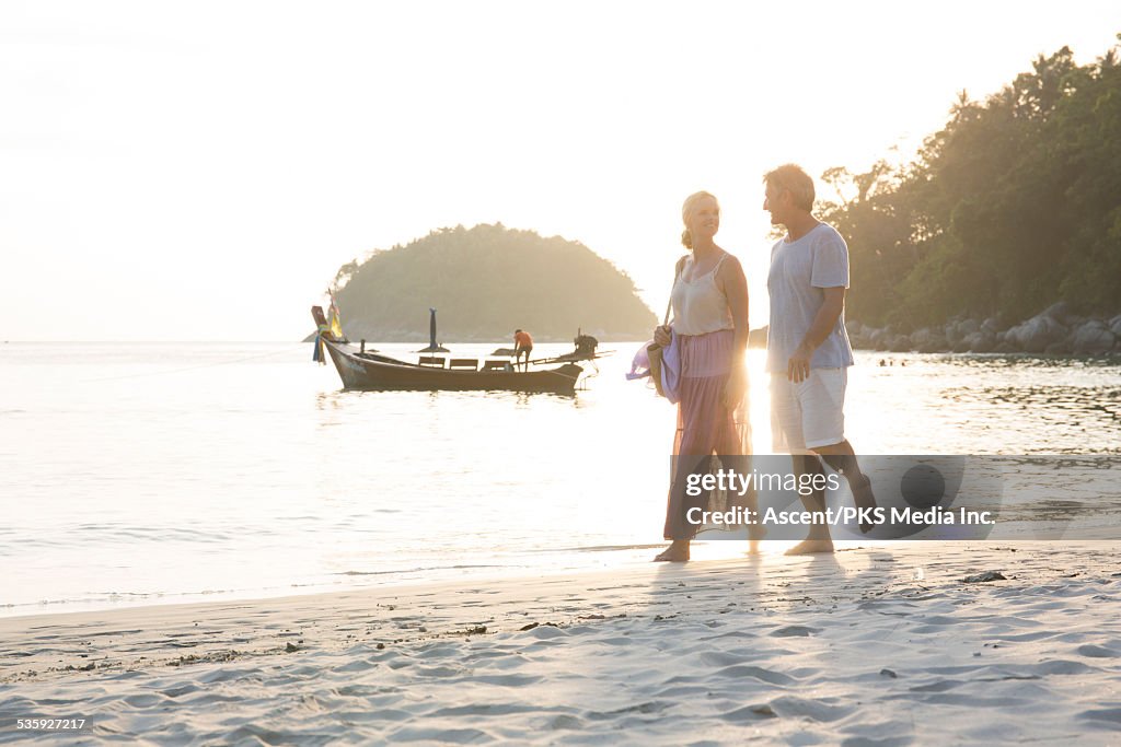 Couple walk along white sand beach, boat behind