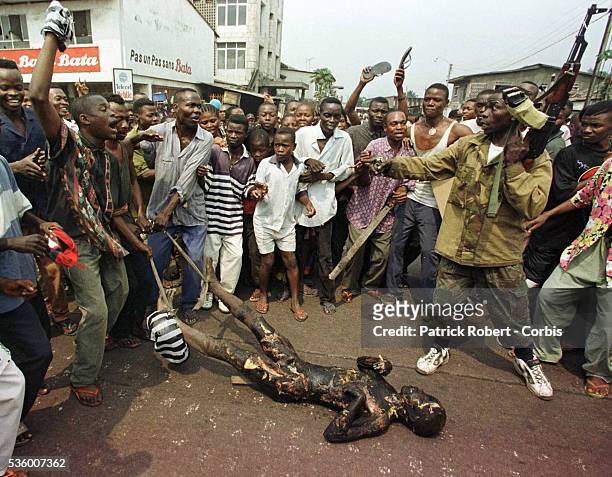 An angry mob drags the body of a burned Tutsi through the streets in front of soldiers.