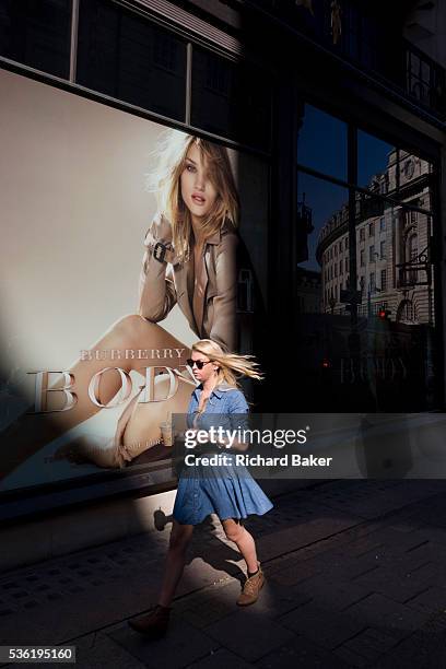 Young woman walks through a pool of sunlight in front of a giant Burberry poster girl ad of the actress Rosie Alice Huntington-Whiteley in central...