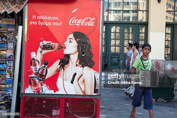 Young boy playing the accordian for money in Monastiraki walks past an aspirational advertisement for Coca Cola. Athens is the capital and largest...