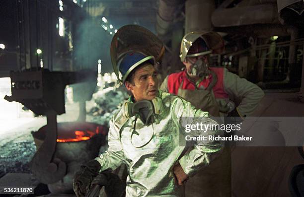 In the heat and dust of a post-communist industrial mill, we see in the foreground two Bulgarian copper manufacturing workers wearing heat-proof...