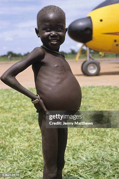 Malnourished child stands in front of a Buffalo plane delivering food aid. Ajiep, Bahr el Ghazal, Sudan. The famine in Sudan in 1998 was a...