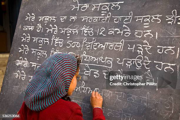 In a Punjabi rural school, a young girl writes on school blackboard whilst other children look on in an open courtyard, India. | Location: Chita...