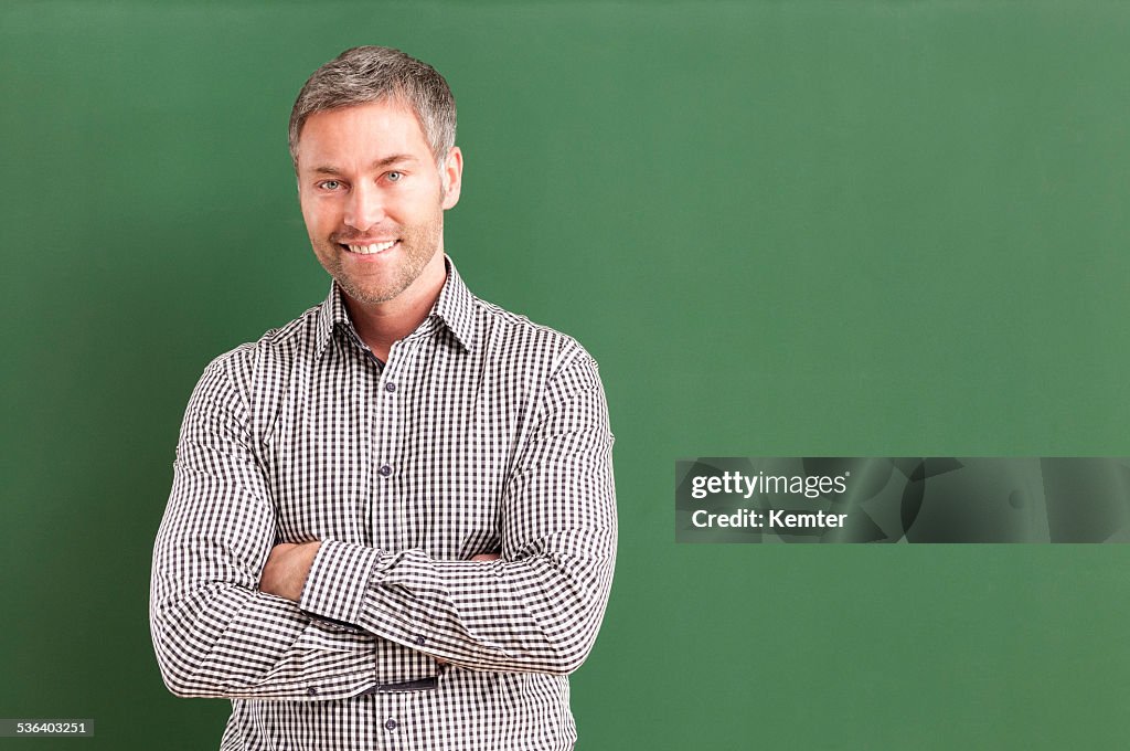 Smiling teacher with arms crossed standing at blackboard