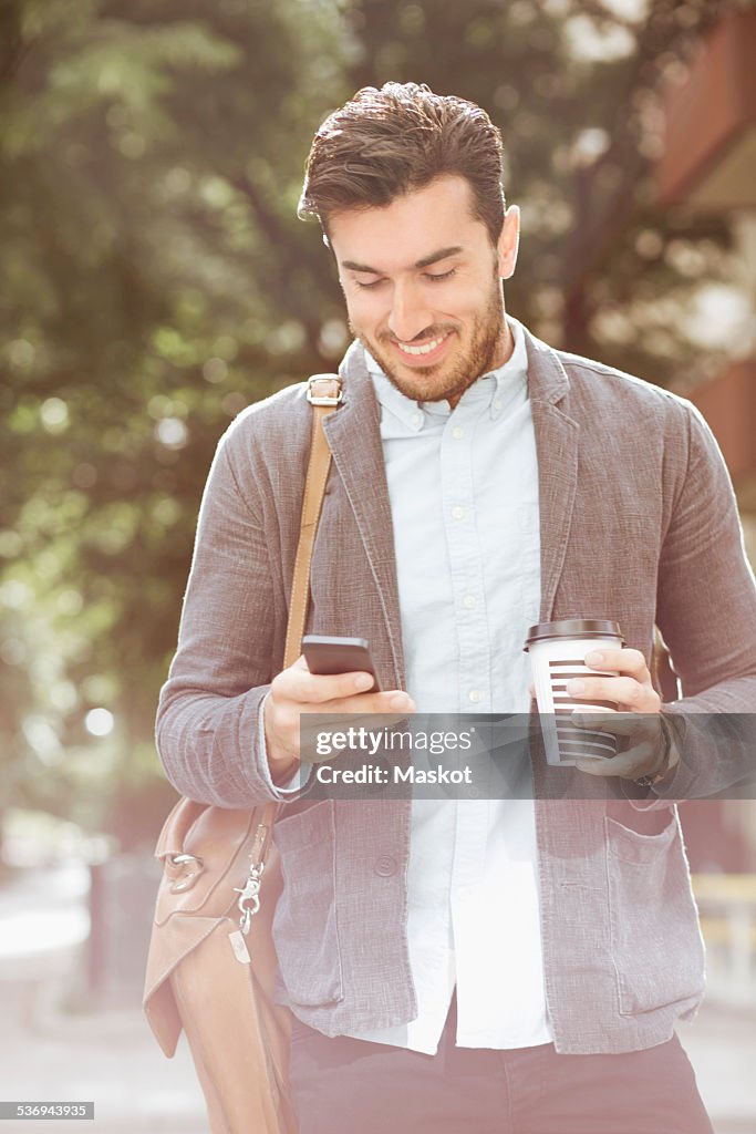 Smiling businessman having coffee while reading text message on smart phone outdoors