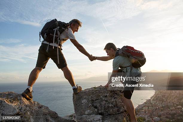 father helps son to pinnacle summit, village below - bergsteiger gipfel stock-fotos und bilder