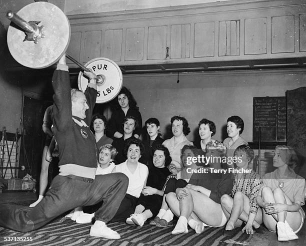 Al Murray, weight lifting coach of the British National team, performing a clean and jerk with a barbell weight in front of a group of office girls,...