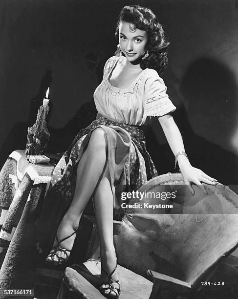 Portrait of actress Rita Moreno, sitting on a table with her feet on a chair, as she appears in the movie 'Cattle Town', 1952.