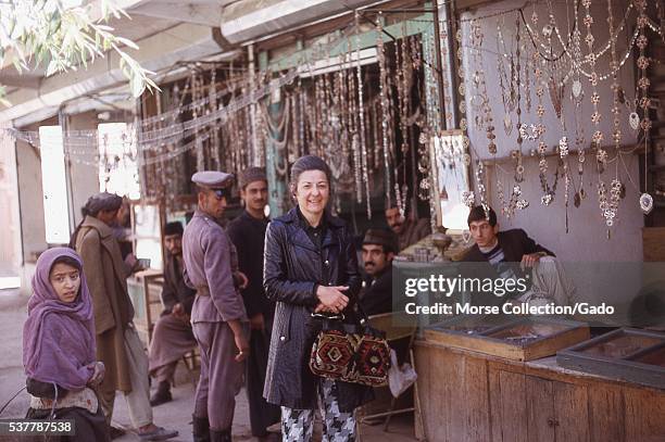 Western woman poses in front of a metalwork jewelry stall at the market in Kabul, Afghanistan, as local Afghanis look on. November, 1973. .