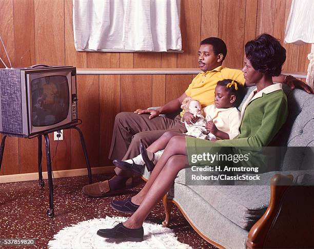 1970s AFRICAN AMERICAN FAMILY FATHER MOTHER DAUGHTER WATCHING TELEVISION SITTING TOGETHER ON SOFA