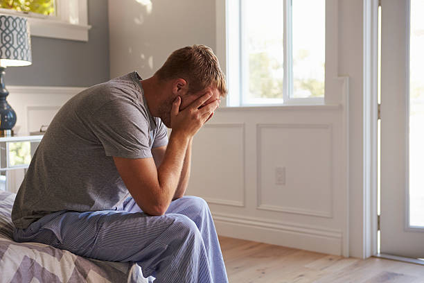 man wearing pajamas sitting on bed with head in hands - depressed man stock pictures, royalty-free photos & images