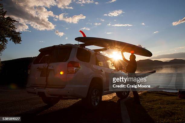 a fit female places her stand up paddle board on a car at sunset on whitefish lake in montana. - lake whitefish stock-fotos und bilder