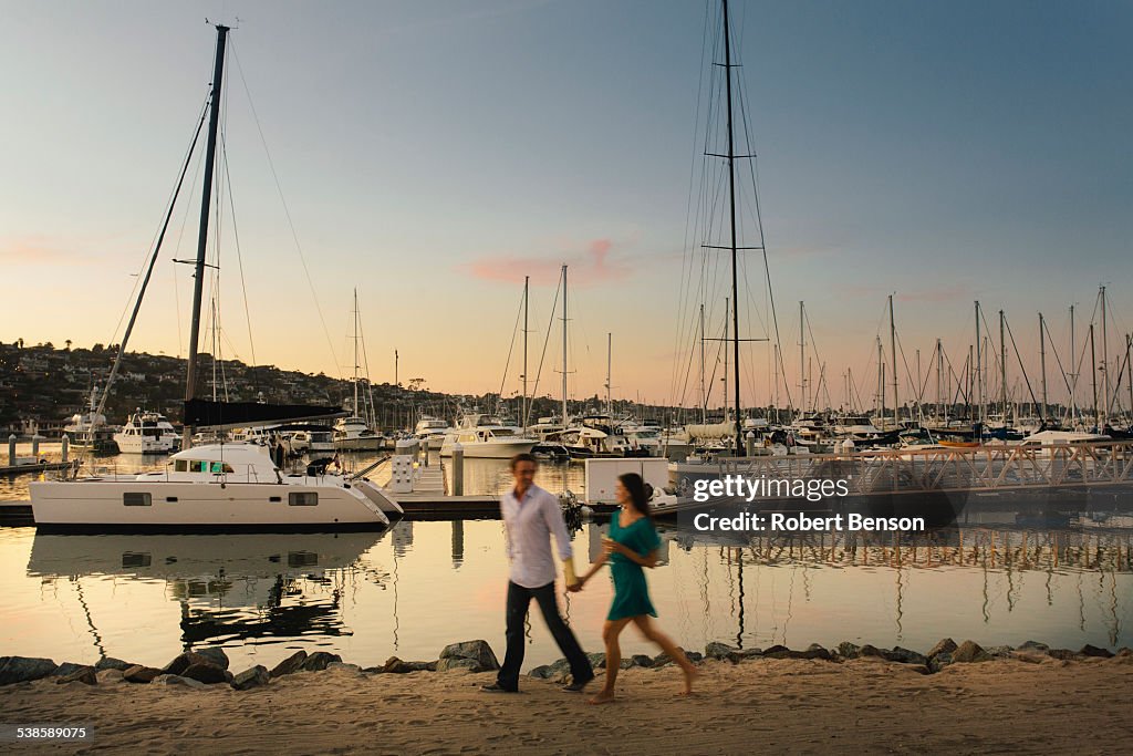 A young girl with a drink in her left hand strolls by the marina along her male partner hand in hand.