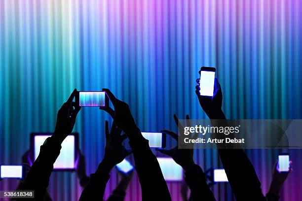 people raise his bright smartphone and tablet device during a night show celebration with dark silhouettes and colorful background. - social crowd stockfoto's en -beelden