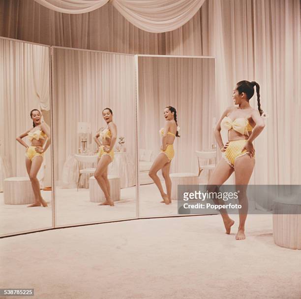 Hong Kong born actress Nancy Kwan pictured wearing a yellow bikini whilst standing in front of three full length mirrors in a dressing room in 1962.