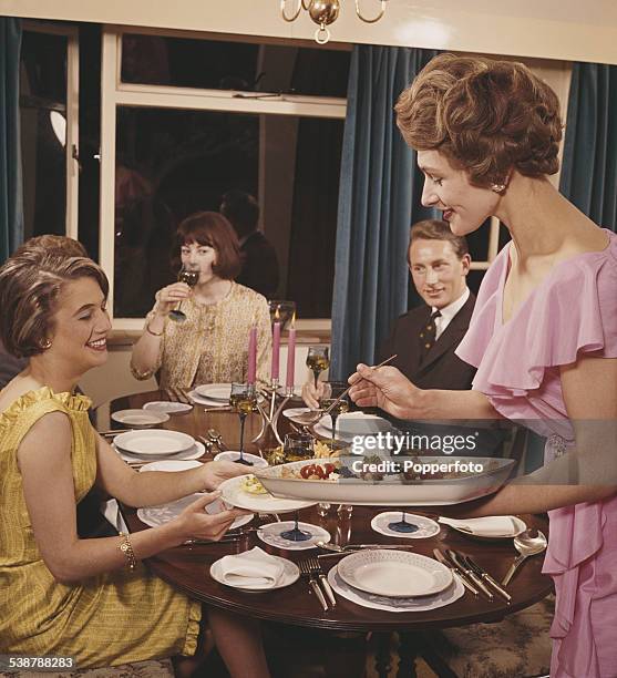 Sixties Fashion - A hostess wearing a pink sleeveless dress serves food to guests at a formal evening dinner party circa 1962.
