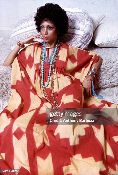 Portrait of an unidentified model, in a red and yellow print dress accented with gold and turquoise jewelry, as she poses on a set of neutral-colored...