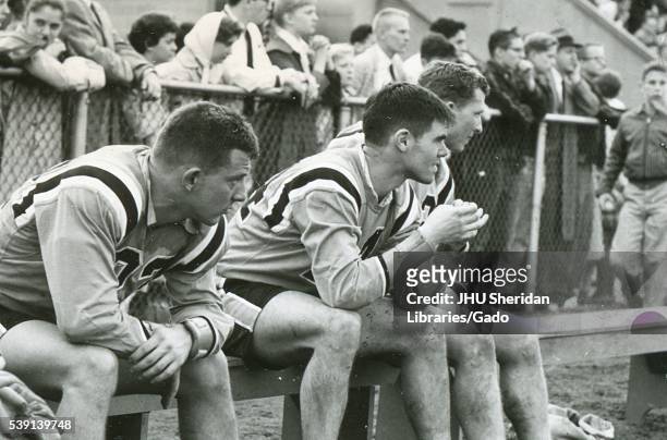 Candid photograph of three male Johns Hopkins University lacrosse players seated on the sideline bench, dressed in uniform, heads turned to the side...