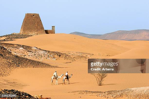 Pyramids of Meroe and two Nubian men dressed in thawbs riding dromedary camels in the Nubian desert of Sudan, North Africa.