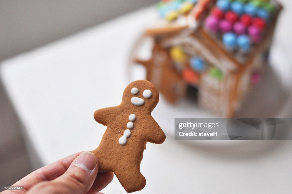 Person's hand holding gingerbread man, gingerbread house in background