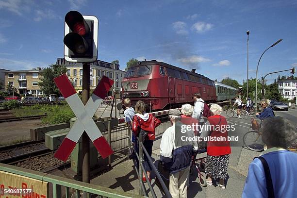 Fußgänger warten hinter einem beschrankten Bahnübergang, den ein Zug passiert. Im Vordergrund eine Ampel mit Andreaskreuz. Bahnschranke; Schranke .